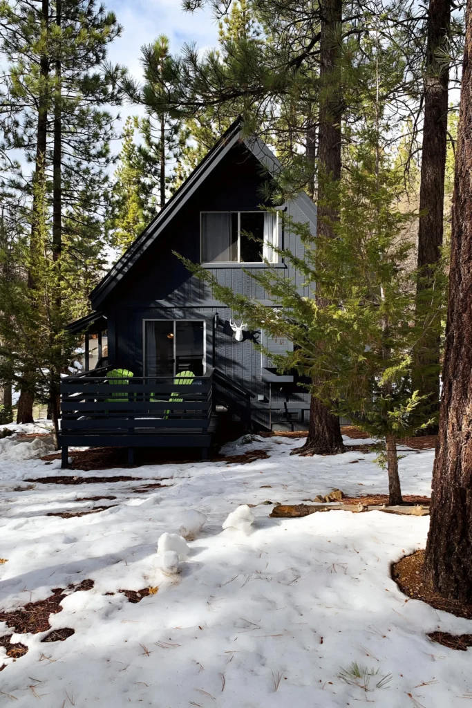 Quaint A-frame cabin surrounded by pine trees and snow, featuring a small front deck with bright green chairs. Modern Cabins