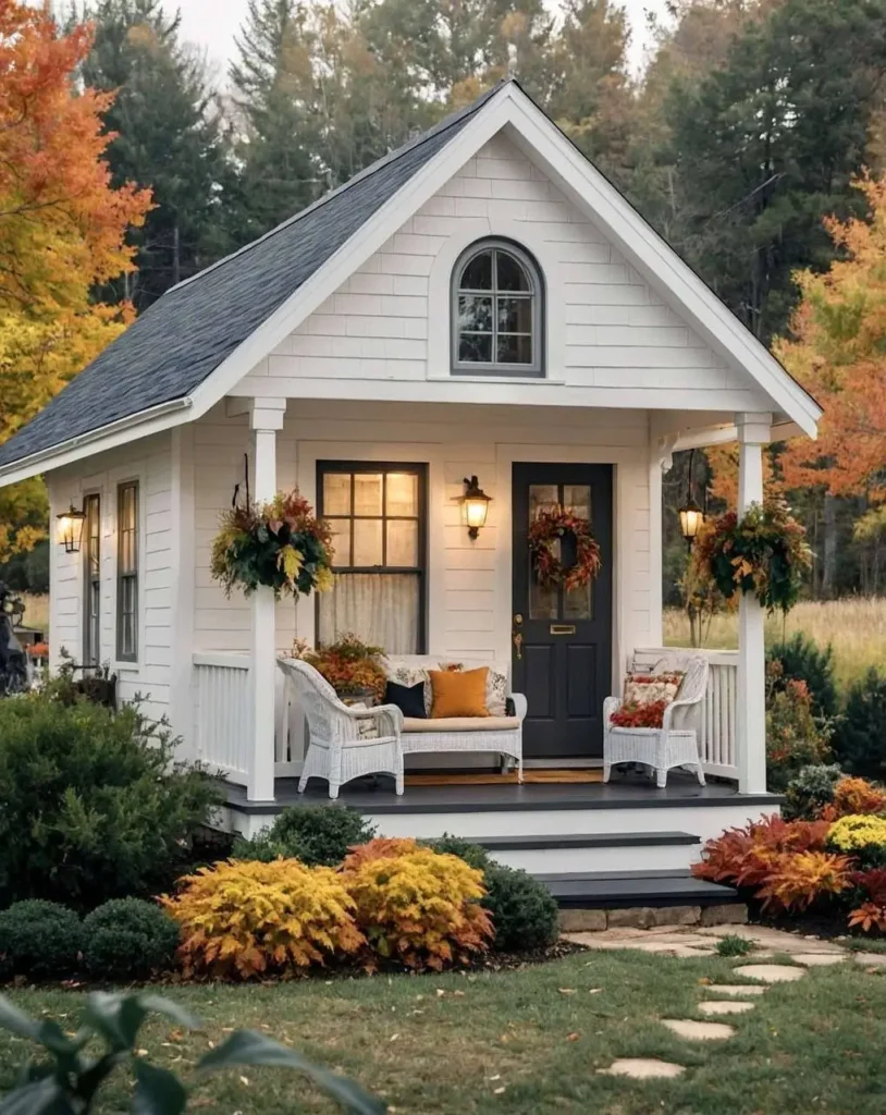 White tiny cottage with a front porch, autumn decor, and colorful fall landscaping surrounded by trees.