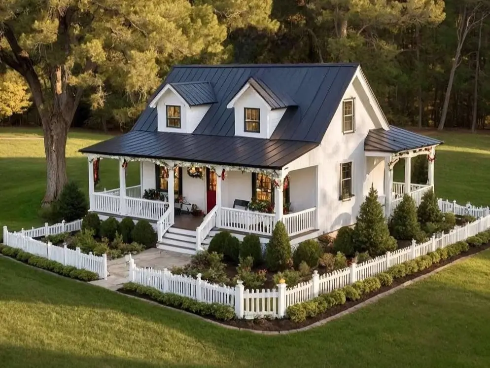 Farmhouse-style tiny house with a black metal roof, white picket fence, wraparound porch, and green lawn.