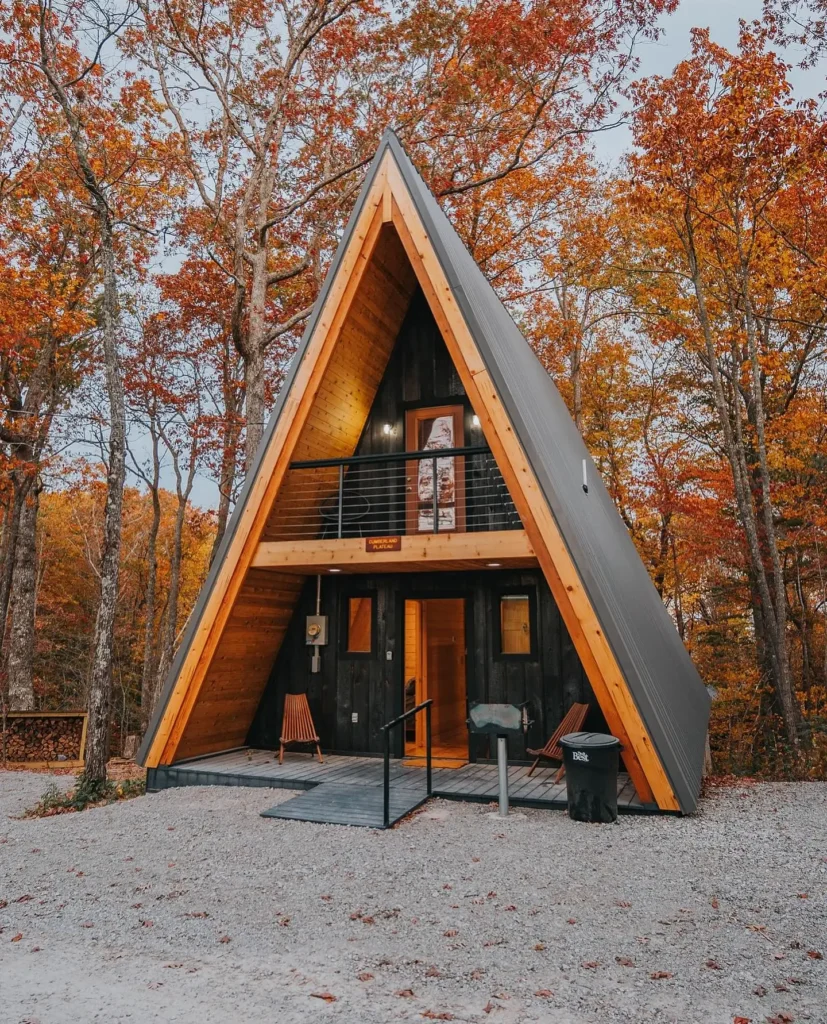 Modern small A-frame house surrounded by autumn foliage, featuring black siding, wooden trim, a second-story balcony, and a ramped entrance.