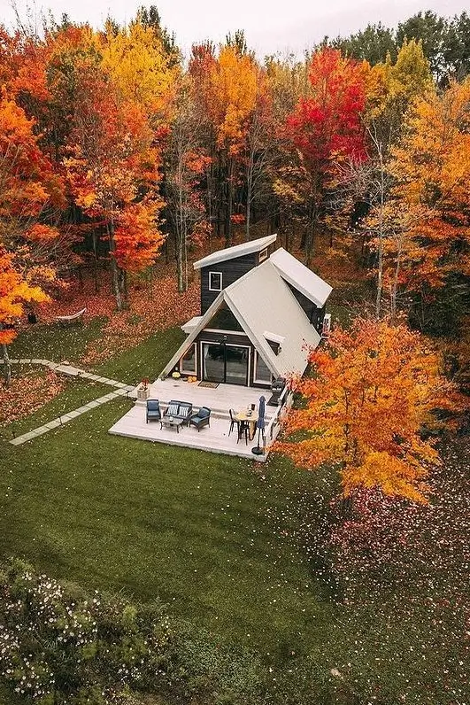 Aerial view of a small A-frame house with an extended patio, surrounded by vibrant autumn trees and a lush green lawn.