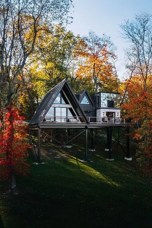 Dual A-frame house elevated on stilts, featuring glass walls, a shared deck, and a modern annex, surrounded by vibrant autumn trees.