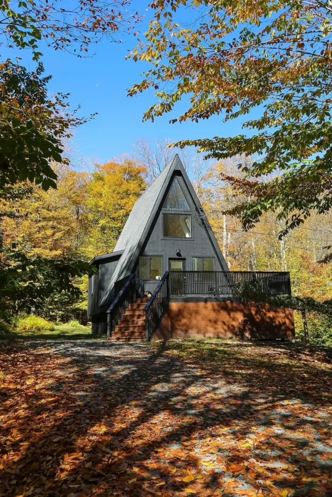 Classic A-frame house with a dark exterior, surrounded by autumn trees, featuring a spacious front deck and wide staircase.