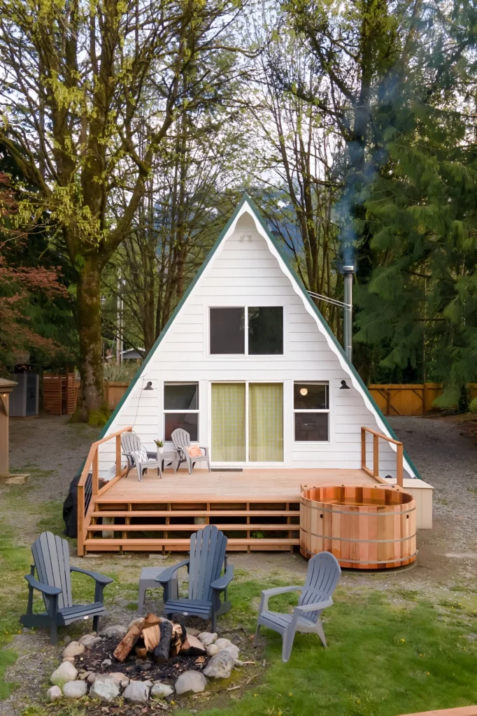 White A-frame cabin with green accents, a front deck, wood-fired hot tub, and a fire pit surrounded by Adirondack chairs in a wooded setting.