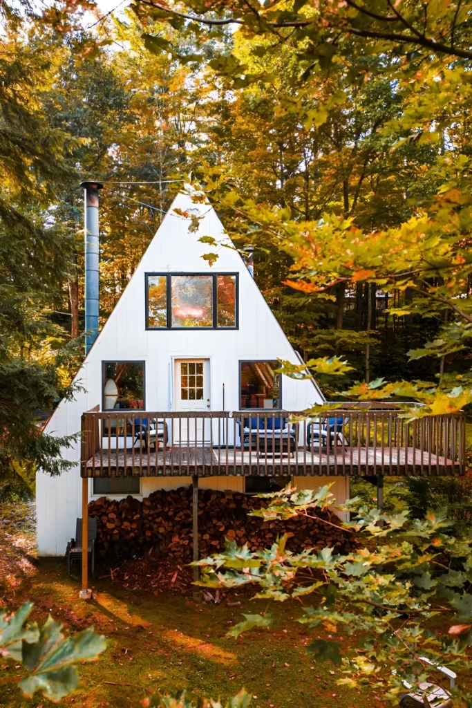 White A-frame cabin with a wraparound deck, large windows, and a stack of firewood below, surrounded by autumn trees.