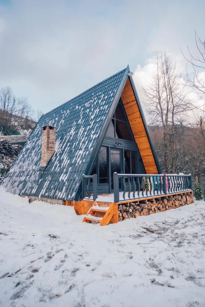 A-frame cabin with a gray roof, wooden accents, a brick chimney, and a front deck surrounded by snow-covered ground.