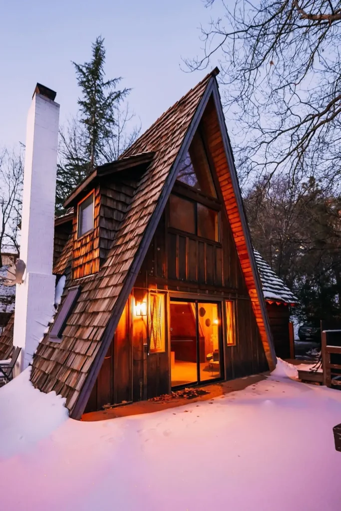 Rustic A-frame cabin with wooden siding, shingle roof, a white chimney, and a warmly lit interior surrounded by snow.