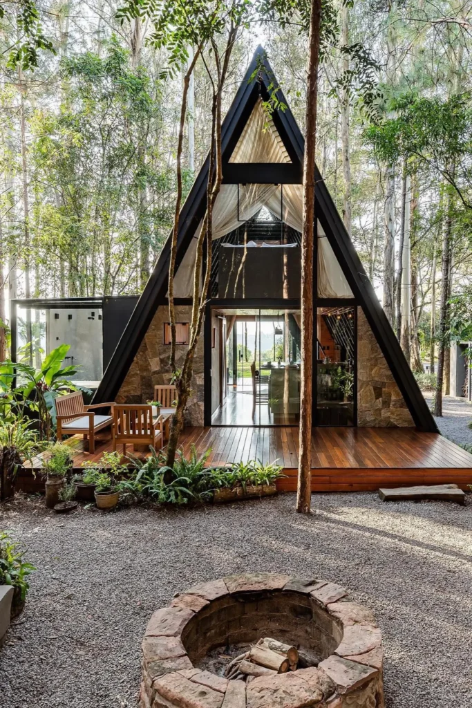 Modern A-frame cabin with stone accents, glass doors, wooden deck, outdoor seating, and a stone fire pit in a forest setting.