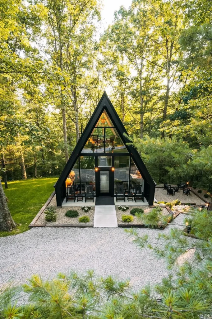 Modern A-frame cabin with floor-to-ceiling glass windows, a landscaped front yard, gravel walkway, and surrounded by green forest.