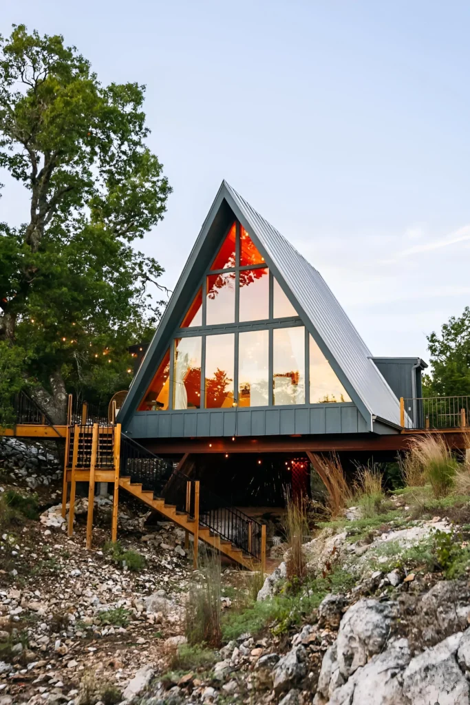 Elevated modern A-frame cabin with large glass windows, a wraparound deck, wooden stairs, and a rocky landscape.
Small A-Frame Houses