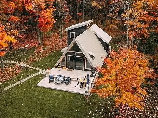 Aerial view of a small A-frame house with an extended patio, surrounded by vibrant autumn trees and a lush green lawn.