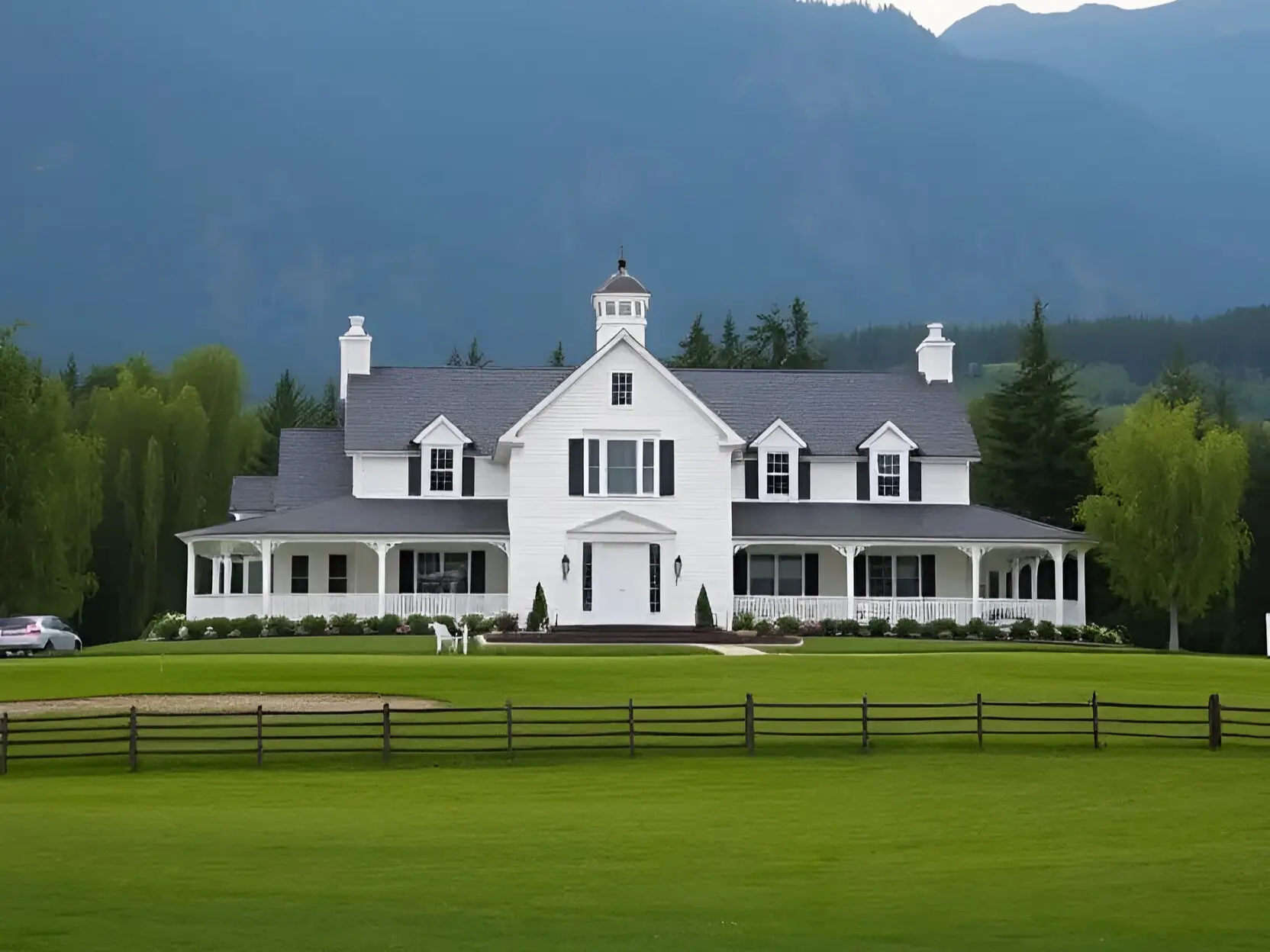 White farmhouse with dual chimneys, wraparound porch, and mountain backdrop during a golden sunset.