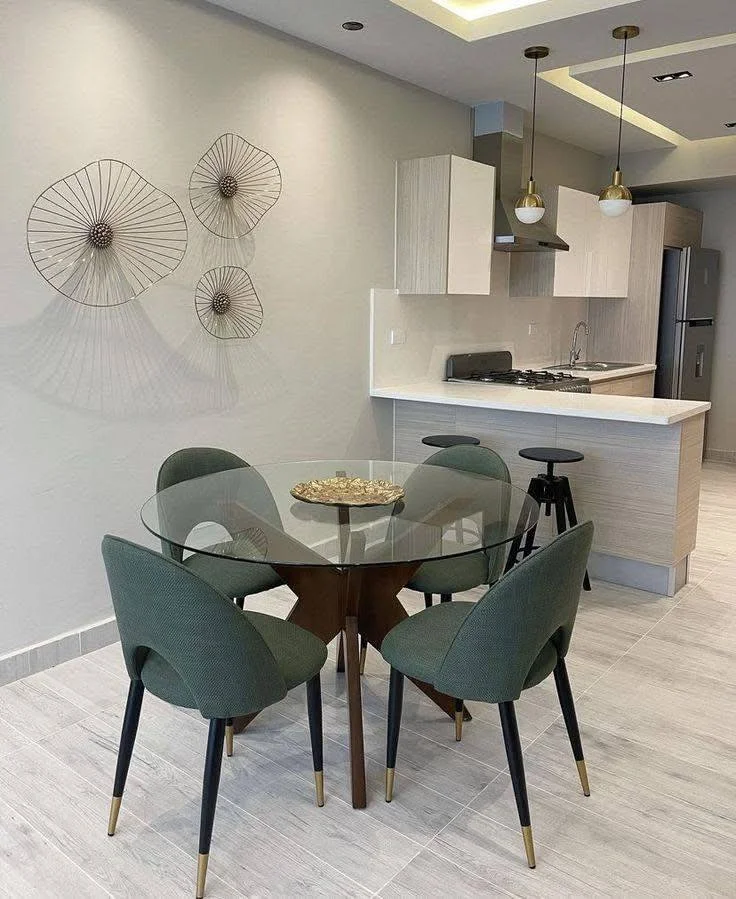 Small dining area with a round glass table, green upholstered chairs, and unique wall decor.