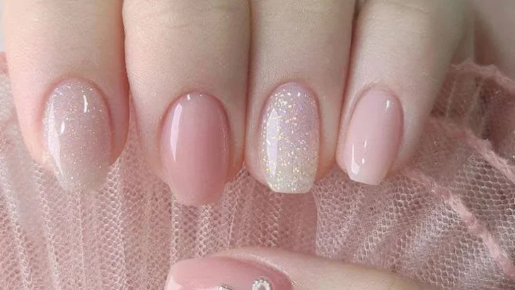 Short pink nude nails with iridescent shimmer and a silver bow charm.