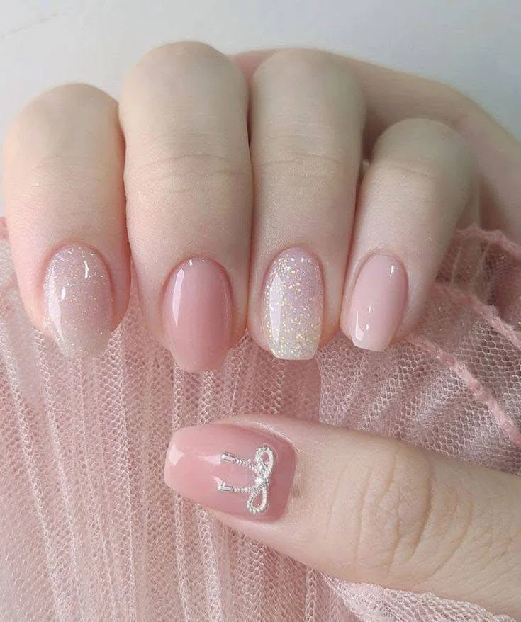 Short pink nude nails with iridescent shimmer and a silver bow charm.