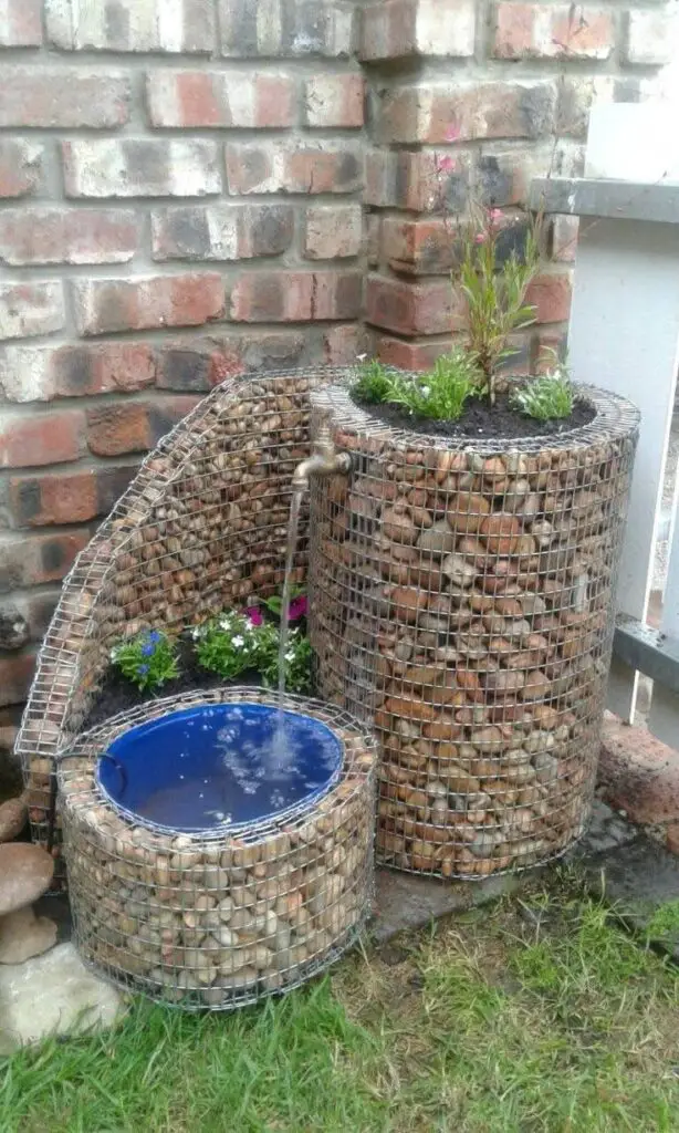 DIY garden water fountain made from river stones encased in wire mesh (gabions), with water flowing from a tap.