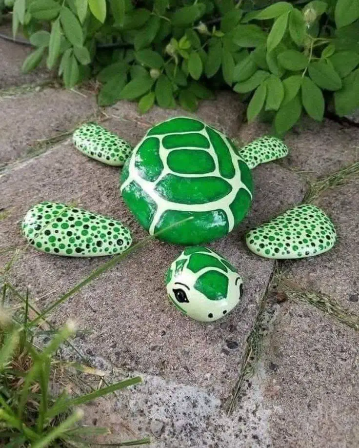 River stones painted to resemble turtles, with green shells and detailed markings, arranged on a stone surface.