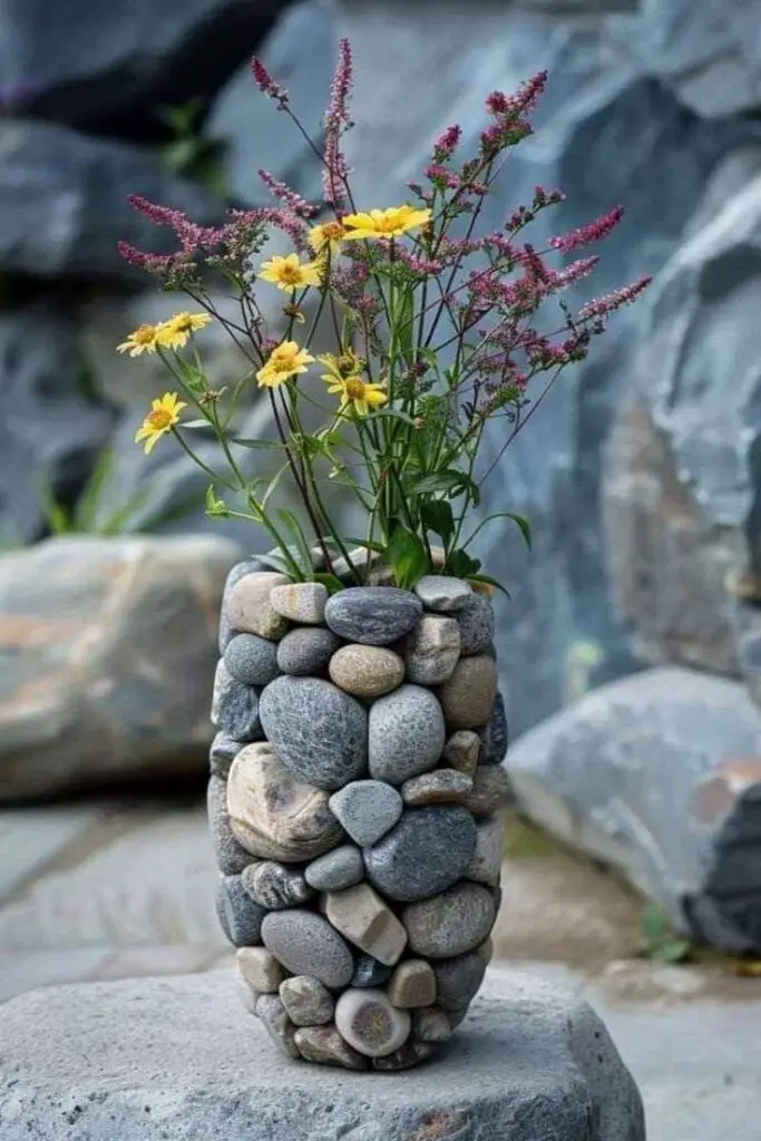 Tall, cylindrical vase made of stacked river stones, holding yellow and purple flowers.