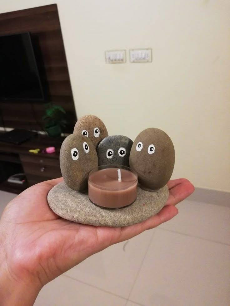 Hand holding a flat river stone with five smaller stones decorated with painted faces, and a tea light candle.