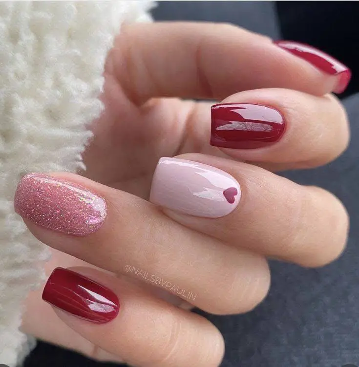 Short square nails with red, pink, and glitter polish, featuring a heart design.