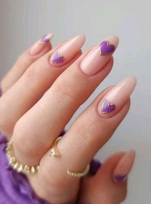 Long almond nails with pale pink polish and purple hearts outlined in gold.