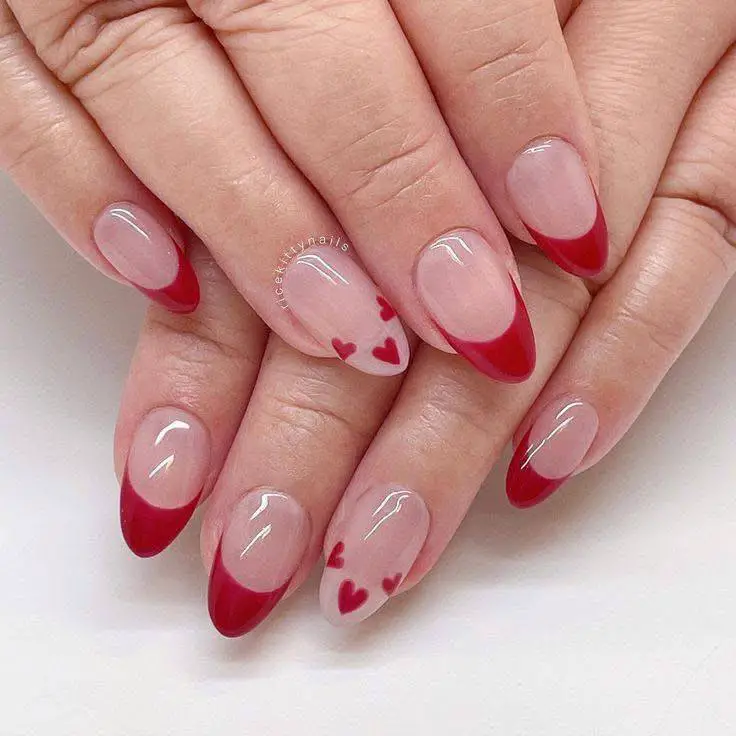 Oval nails with a sheer pink base, deep red French tips, and small red heart accents.