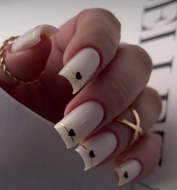 Long square nails with creamy white polish, gold stripes, and small black hearts.