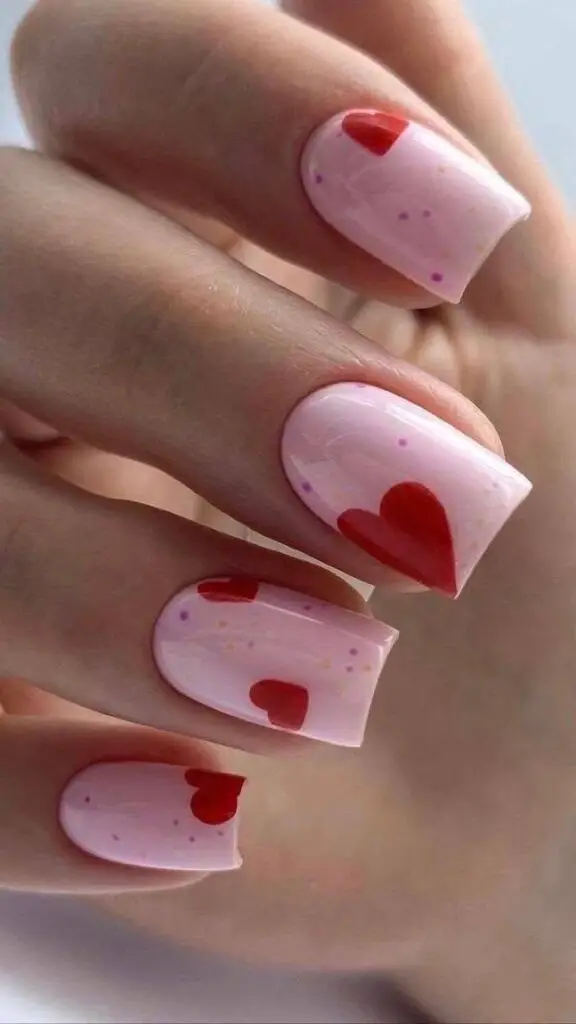 Medium square nails with glossy light pink polish and varied-size red heart accents.