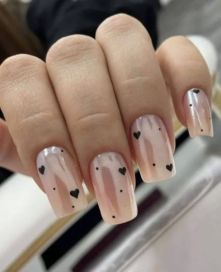 Square nails with iridescent light pink polish and small black hearts and dots.