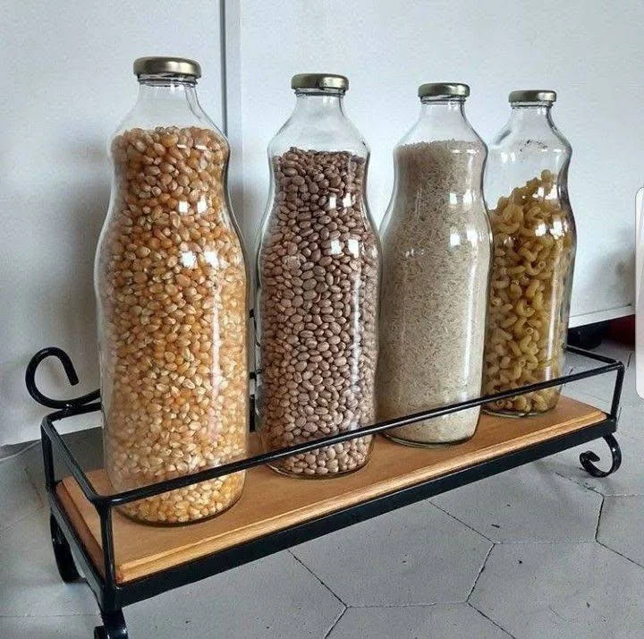 Four glass bottles filled with dry goods (corn, beans, rice, pasta) on a decorative shelf.