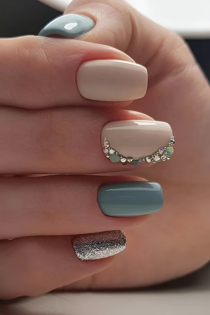 Short nails with teal, beige, and silver glitter polish, featuring a rhinestone arc design.