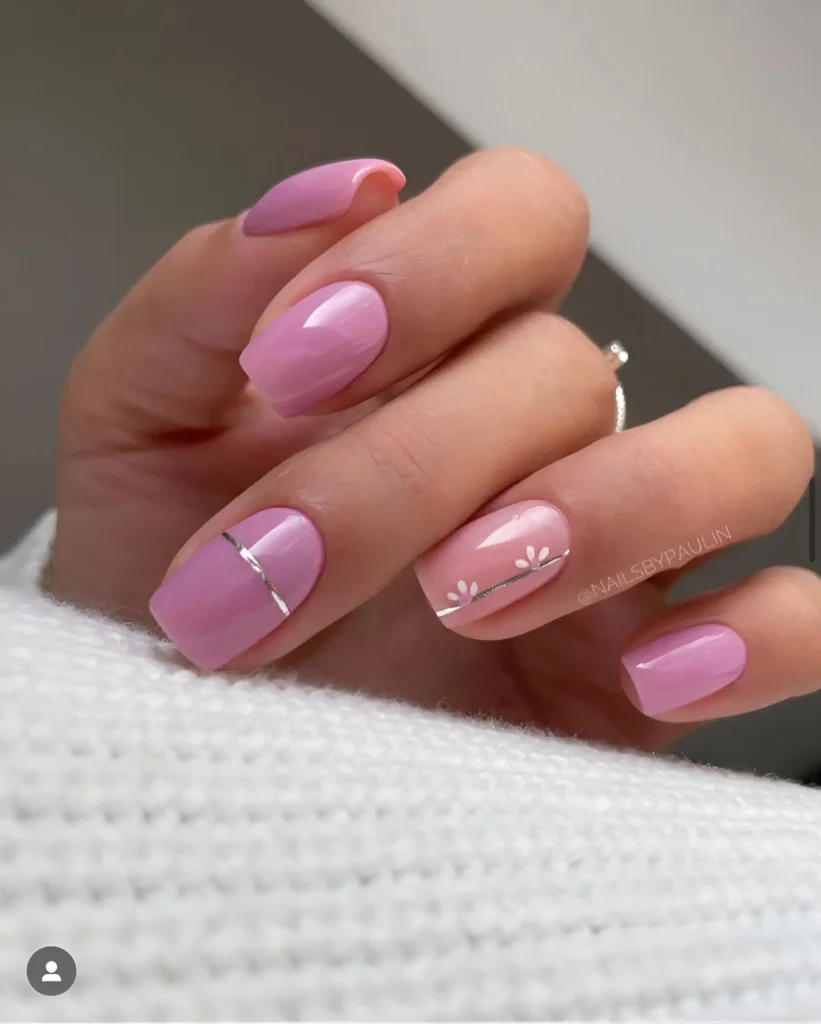 Short, rounded nails. Some are painted solid shades of pink, ranging from light to medium. Two nails feature a pale pink base with small, hand-painted white flowers and a thin, vertical silver stripe.