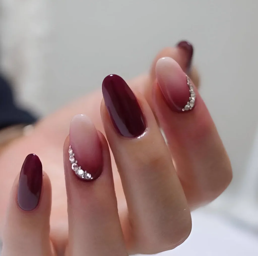 Long, oval-shaped nails. Some nails are a solid, glossy burgundy. Others feature a gradient, blending from a nude shade at the base to burgundy at the tip, with a crescent-shaped line of small crystals along the cuticle line.