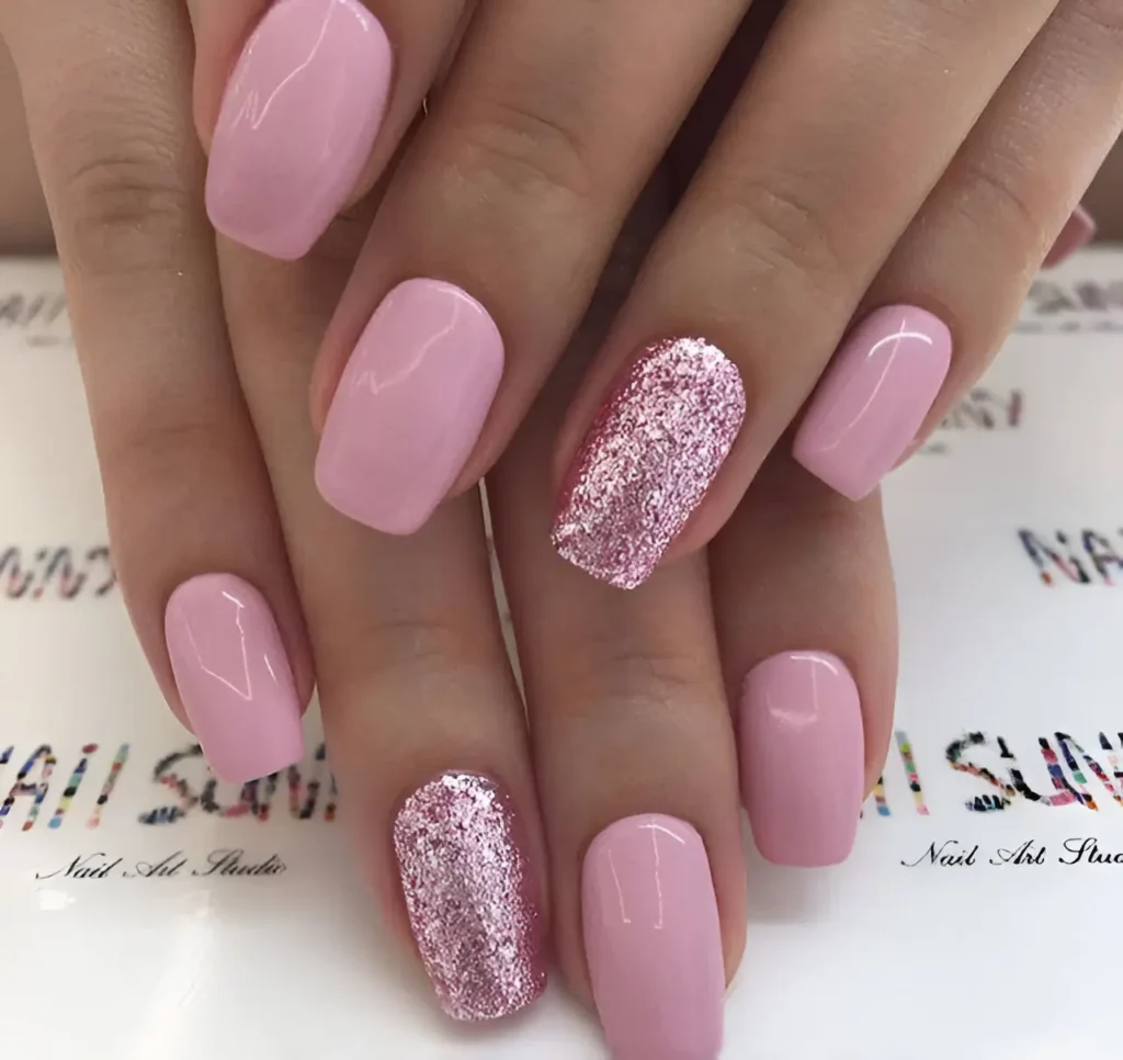 Short, rounded nails. Most are painted a solid, glossy, light pink. One nail on each hand is fully covered in a light pink glitter.