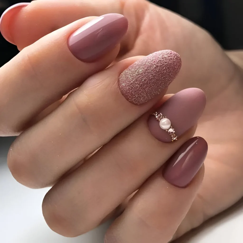 Almond-shaped nails. Some are painted in solid, glossy shades of rose, ranging from light to medium. One nail is covered in a textured, matte rose-gold glitter, and one features a row of small pearls and gold beads along the base.