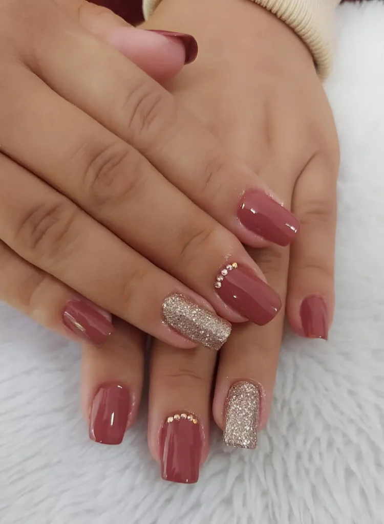 Square-shaped acrylic nails. Most are painted a solid, glossy deep rose. One nail on each hand is covered in champagne-colored glitter, with a line of small crystals along the base of the nail.