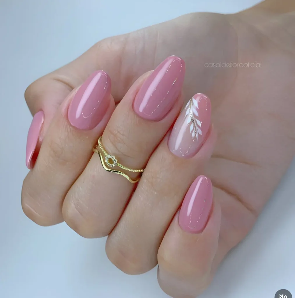 Almond-shaped nails. Most are painted a solid, glossy pink. One nail has a nude base with a hand-painted white design resembling leaves or feathers, with a few small, clear crystals.