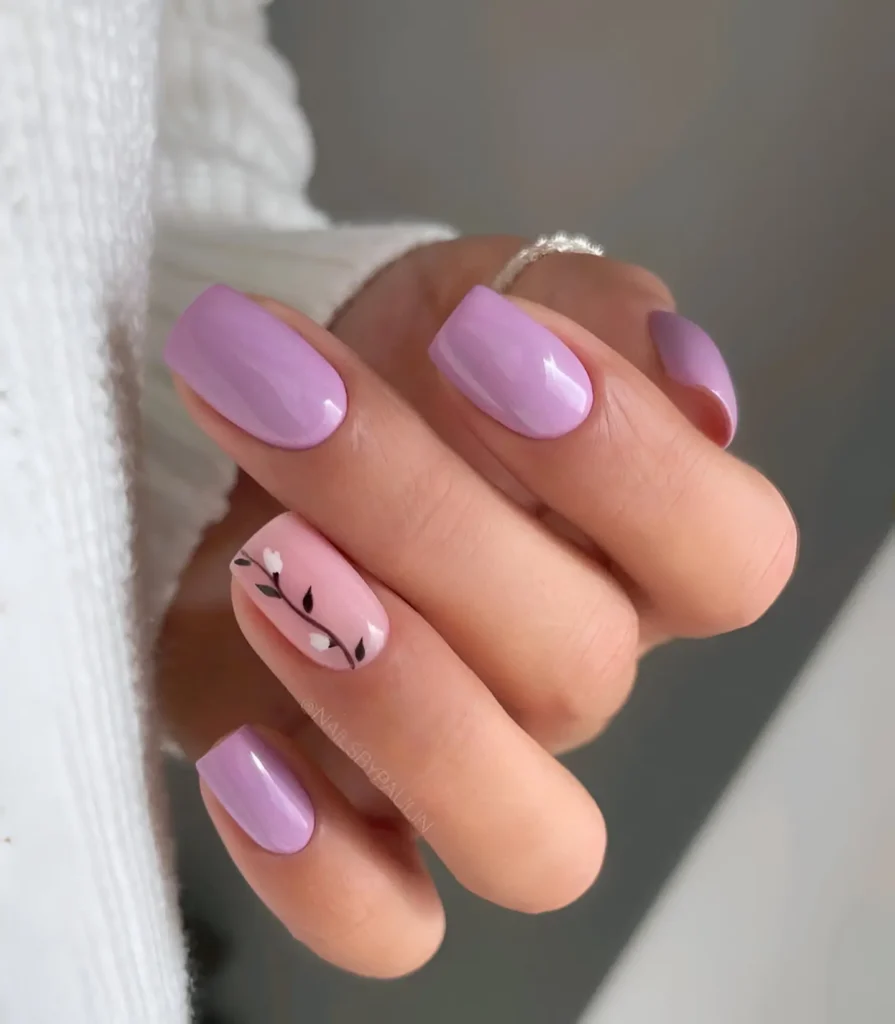 Short, rounded nails. Most are painted a solid, glossy lavender. One nail has a pale pink base with a hand-painted floral design featuring thin black stems and small white flowers.