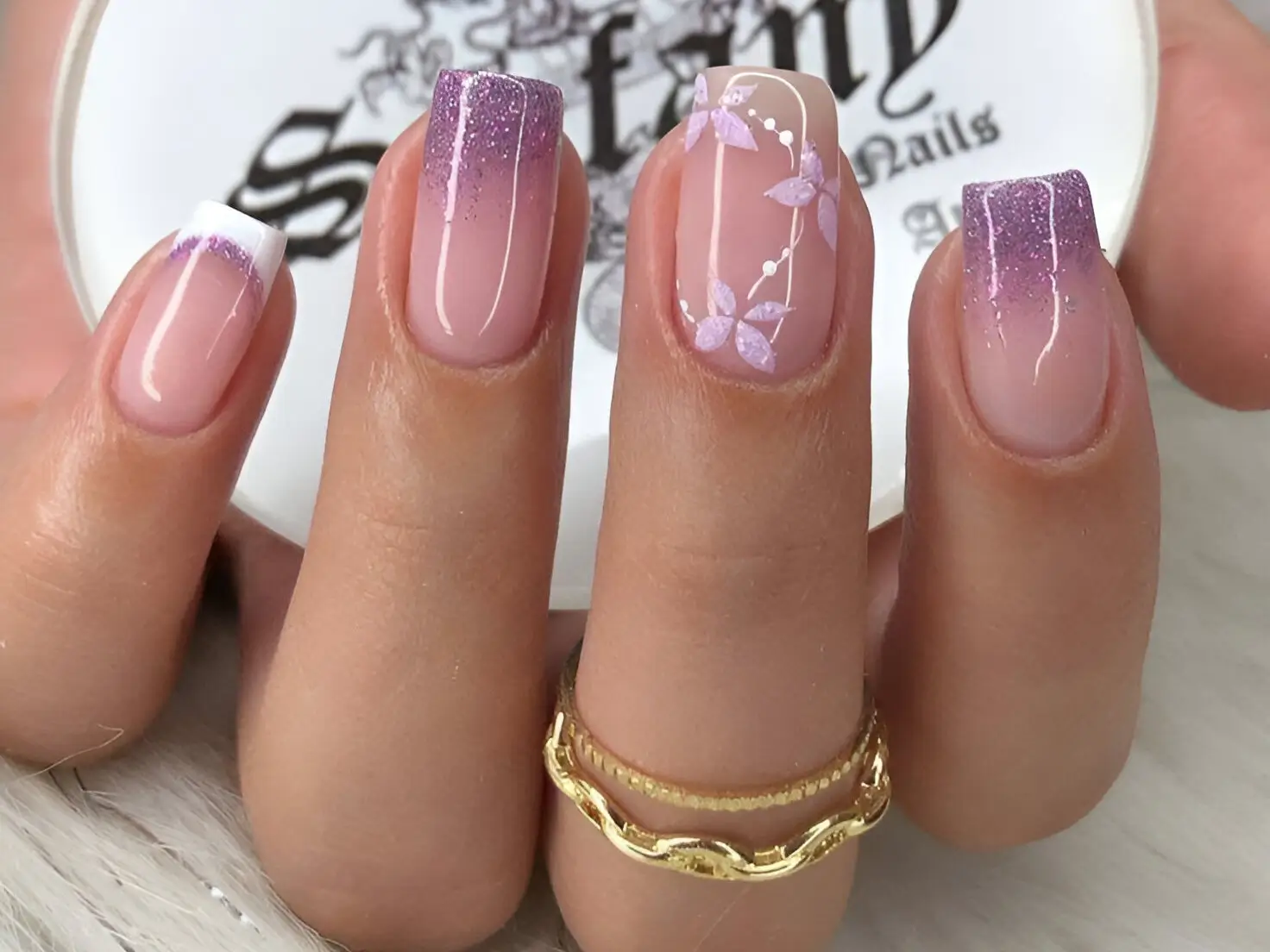 Square-shaped nails with multiple designs. One nail has a classic French tip, two have a gradient from clear at the base to lavender glitter at the tip, and one features hand-painted lavender flowers and leaves on a clear base.