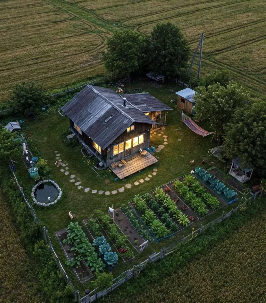 Aerial view of a rustic off-grid cabin with a vegetable garden, pond, and warm glowing lights at dusk.