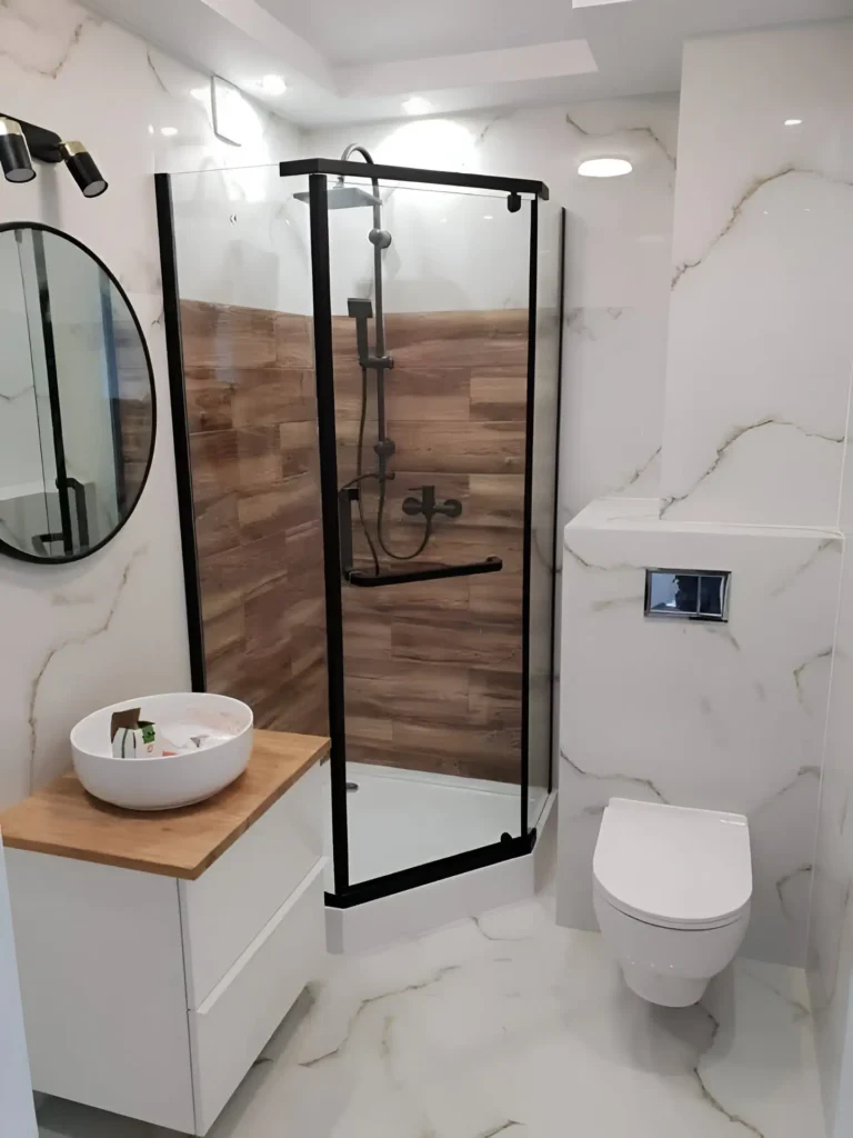 Modern small bathroom with marble-look tile, wood-look shower accent, black framed shower, and round mirror.