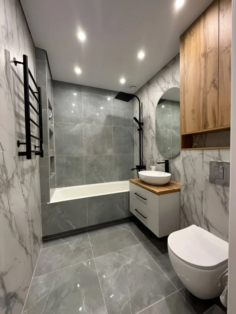 Modern small bathroom with bathtub-shower combination, gray and marble-look tiles, black fixtures, and wall-mounted toilet.