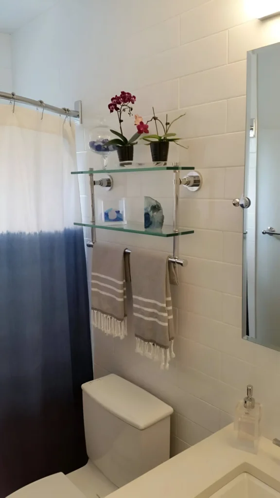 Two-tiered glass shelf with chrome supports, mounted above a toilet; the lower tier acts as a towel rack, holding two folded towels, while the upper tier displays small plants and decorative items.