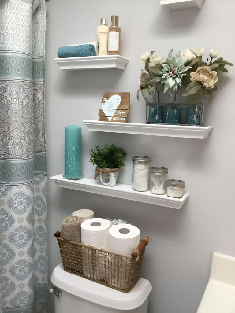 Multiple white shelves of varying lengths mounted above a toilet, holding rolled towels, toilet paper in a basket, decorative items, and toiletries; a sign reading "Love Lives Here" is displayed.