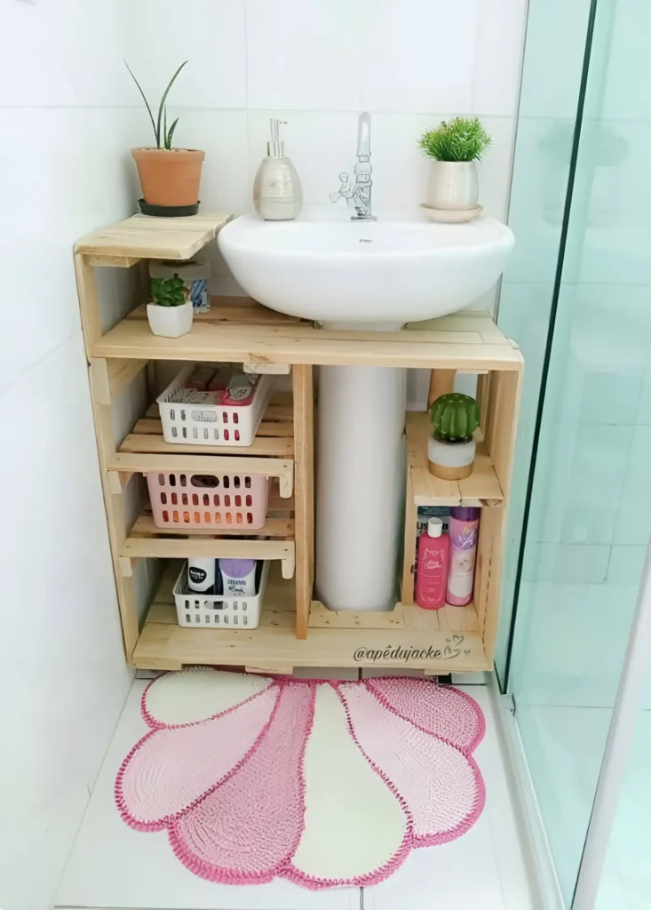 Pedestal sink with a custom-built, light wood shelving unit underneath, creating open storage; small baskets and containers hold toiletries, and small plants are placed on the top and sides.