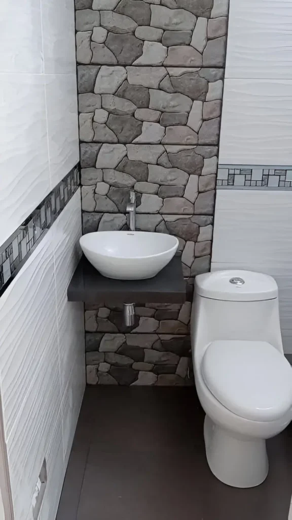 Small bathroom with a wall-mounted gray floating vanity topped with a white vessel sink; a stone-patterned accent wall is behind the vanity, and a white toilet is next to it.