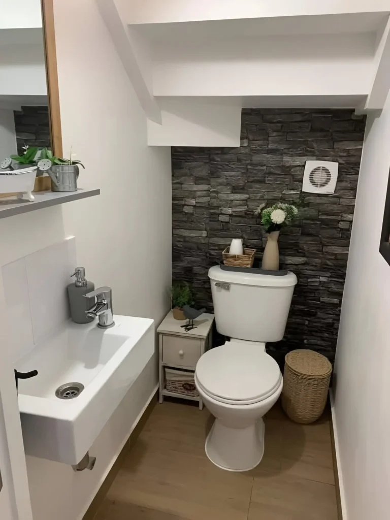 Small bathroom with a tiny white cabinet with drawers placed between a wall-mounted sink and a toilet; a stone-patterned accent wall is behind the toilet, and a woven basket sits beside it.