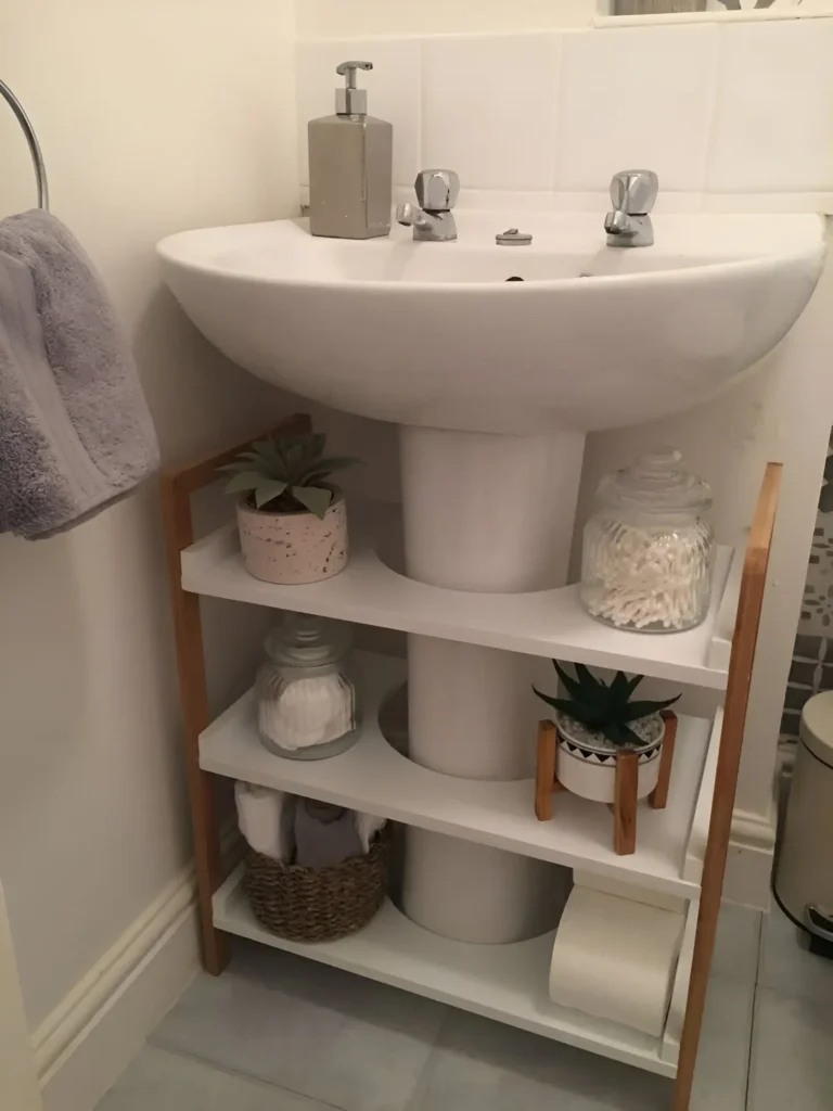 Three-tiered white and light wood ladder shelf placed under a pedestal sink; shelves hold small plants, jars of cotton balls, and a basket with washcloths.