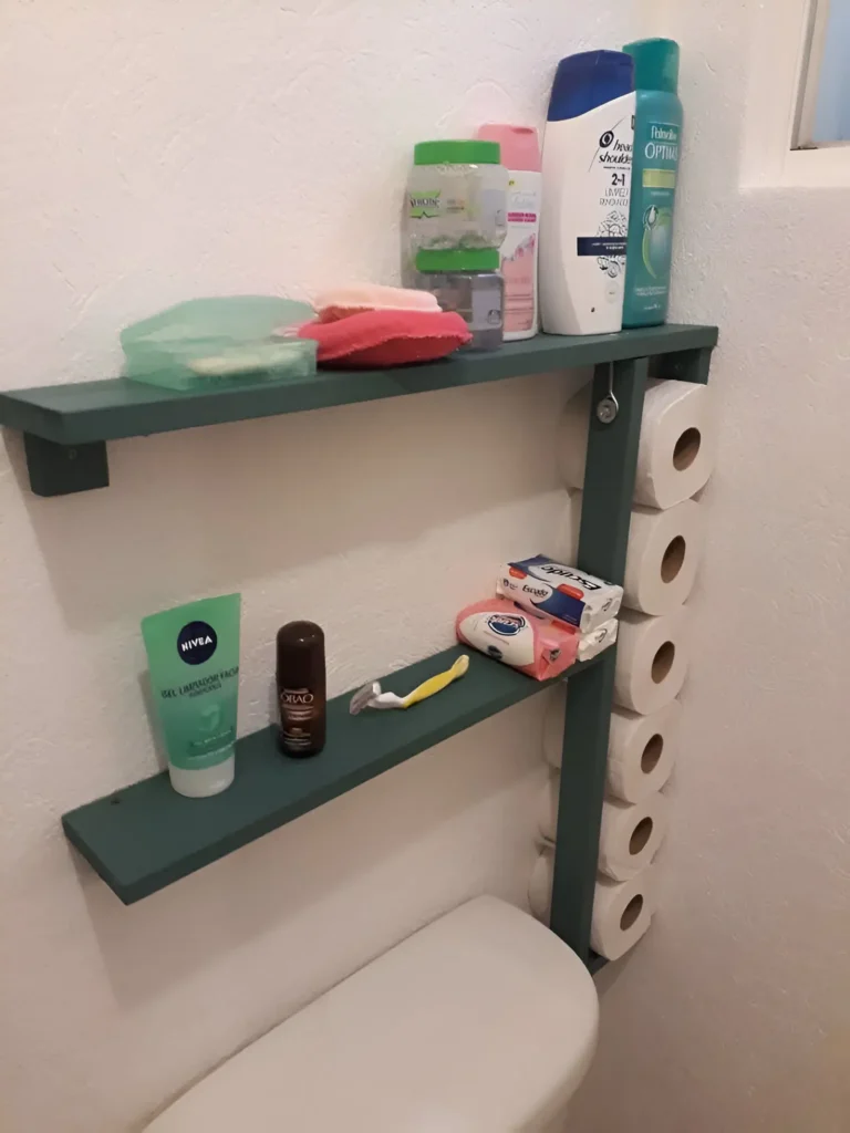 Teal-colored wooden shelf unit mounted beside a toilet; one section holds multiple toilet paper rolls vertically, and two shelves above hold toiletries, soap, and a razor.