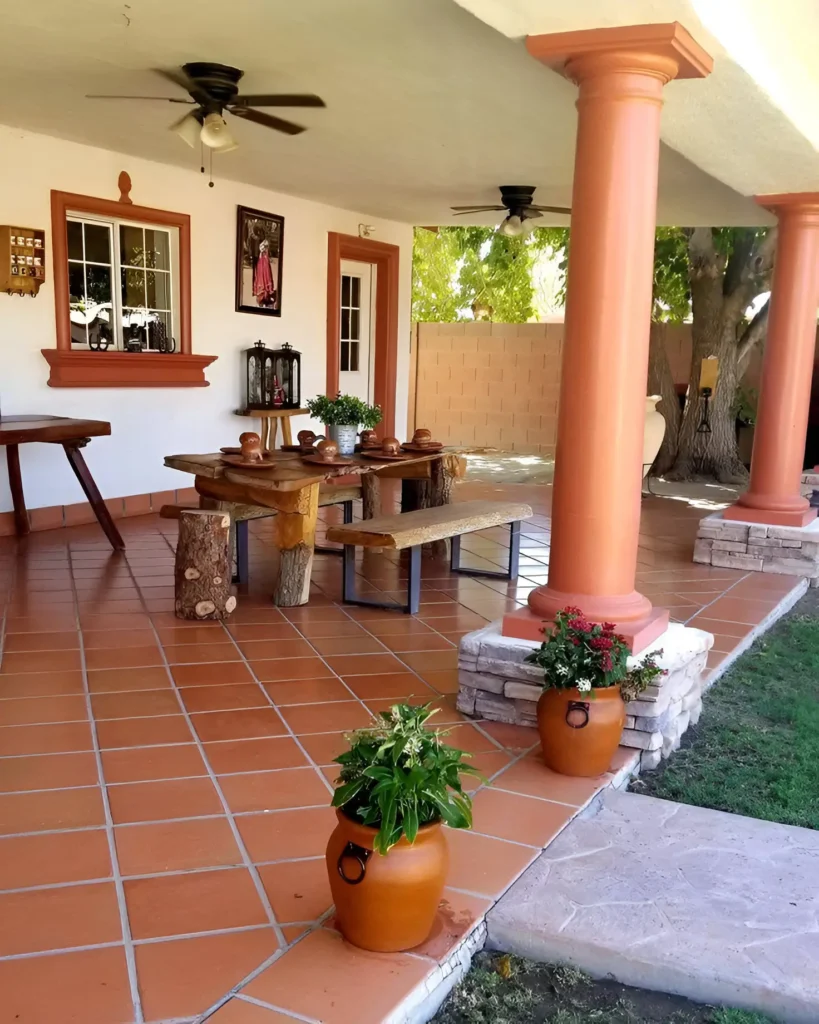 Outdoor patio with terracotta floor tiles, orange pillars, matching orange potted plants, and wooden dining furniture.